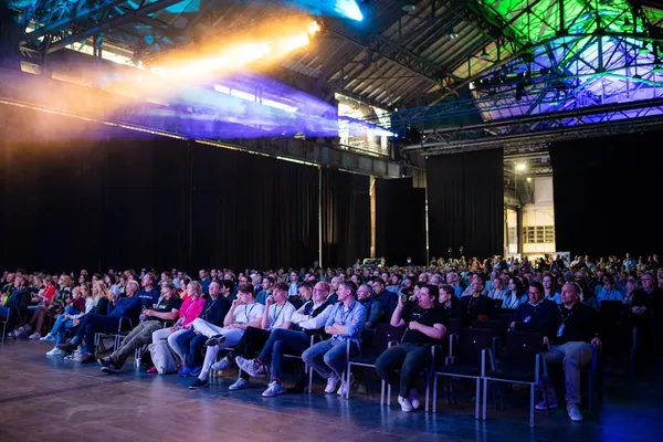 „Publikum sitzt in Stuhlreihen bei einer Konferenz in einer großen, dunkel beleuchteten Industriehalle.“